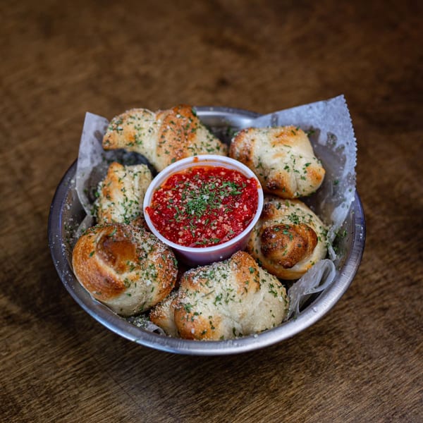 Garlic knots with parmesan and parsley, served in a metal bowl with a side of marinara sauce.